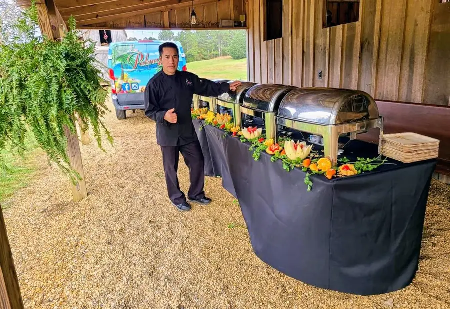 A Las Palmas catering grill and buffet station set up outdoors at a rustic barn venue, attended by a team member