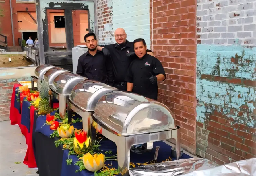 Las Palmas catering staff in black uniforms standing behind a line of chafing dishes with carved-fruit garnishes at a brick-walled venue