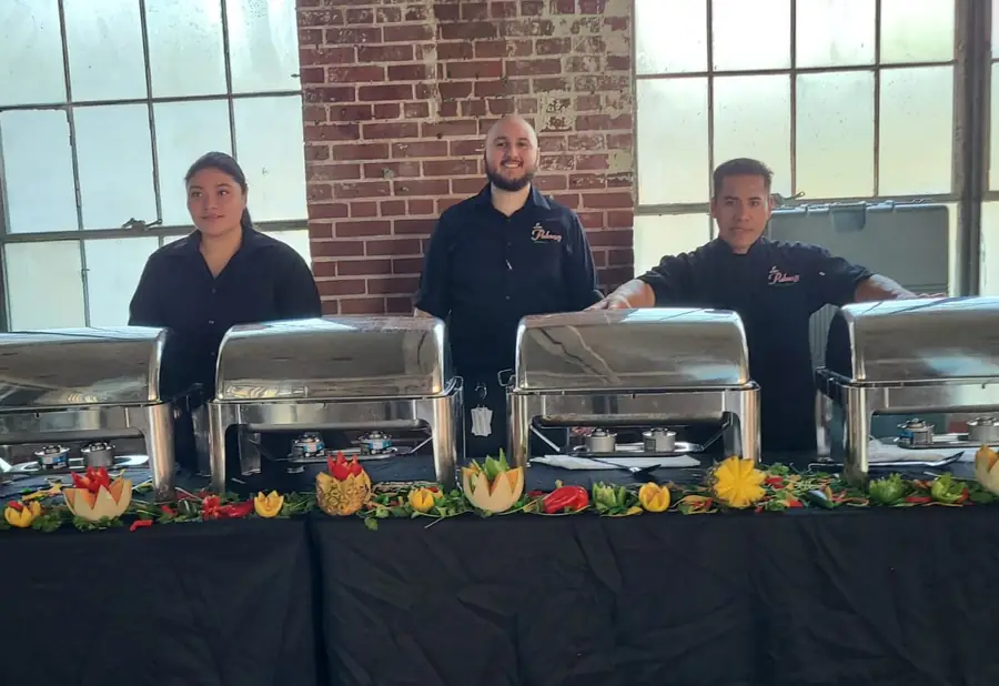 Three Las Palmas team members behind a full catering spread of chafing dishes garnished with carved pineapple and peppers indoors